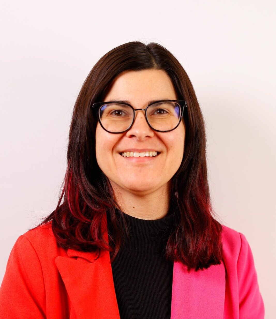 Stephanie Birmingham with brown hair and glasses wears a black shirt with a bright pink and red blazer.