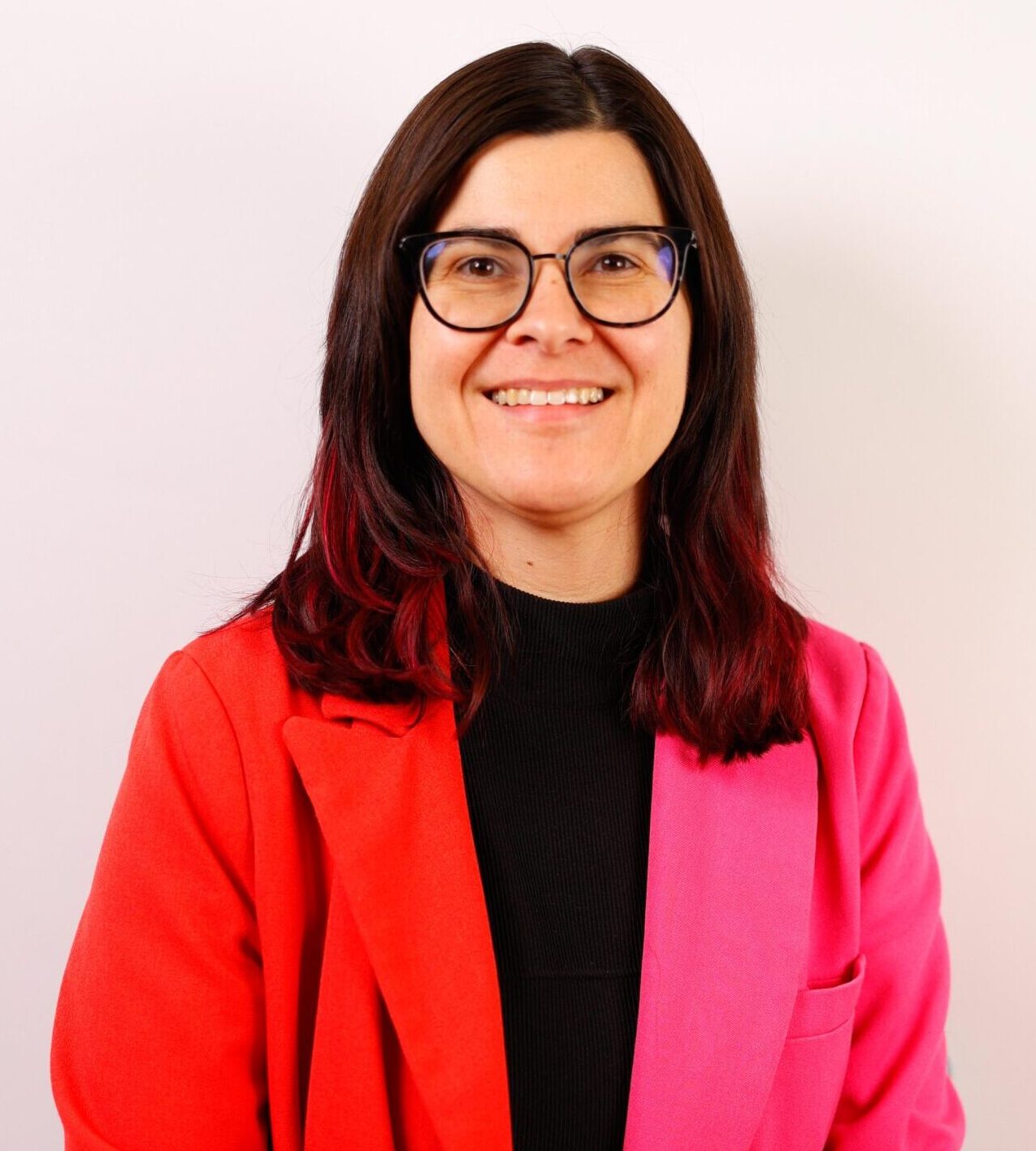 Stephanie Birmingham with brown hair and glasses wears a black shirt with a bright pink and red blazer.