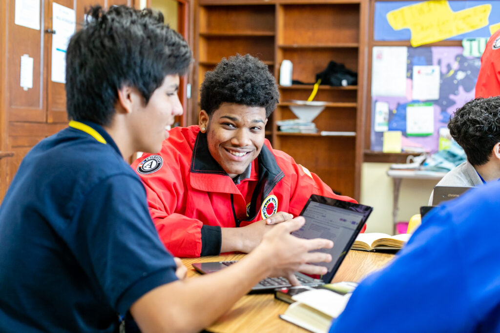 CYNY AmeriCorps Member smiles and sits with a student in front of a laptop doing schoolwork