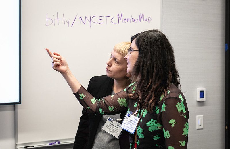 Two women point at a projector in a classroom space.