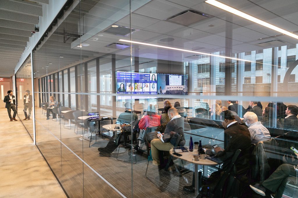 A classroom with glass walls and attendees seated facing a panel of speakers.