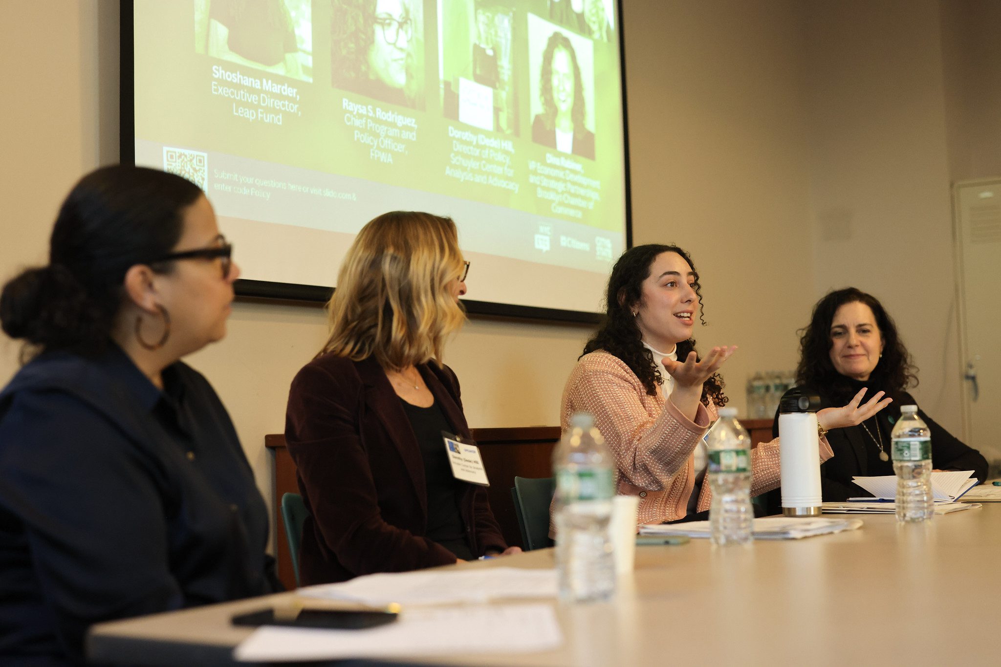 For women sit at a table with water bottles on it in front of a projector.