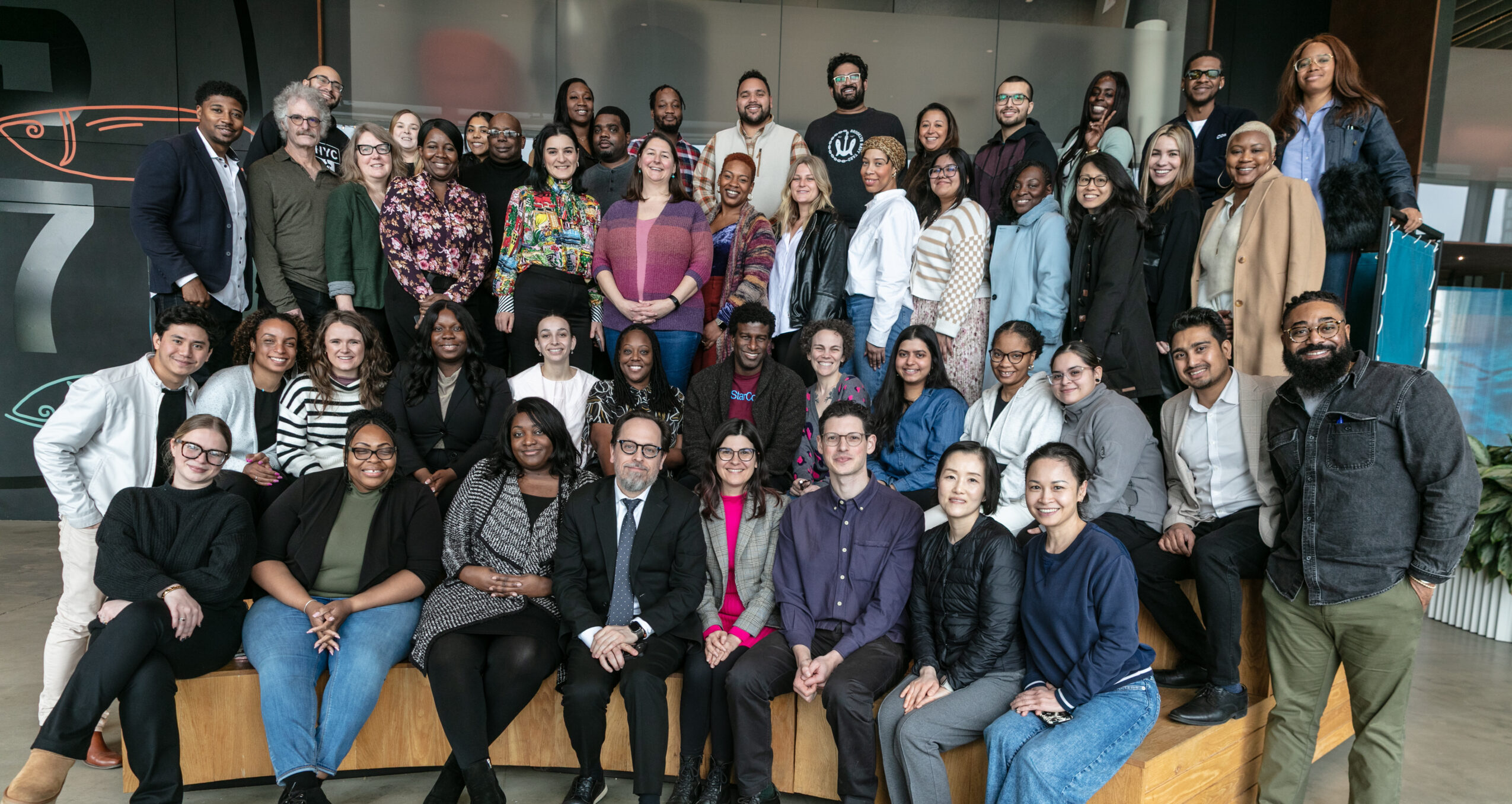 A group of 70 professionals on wooden risers at Pier 57 in Manhattan.