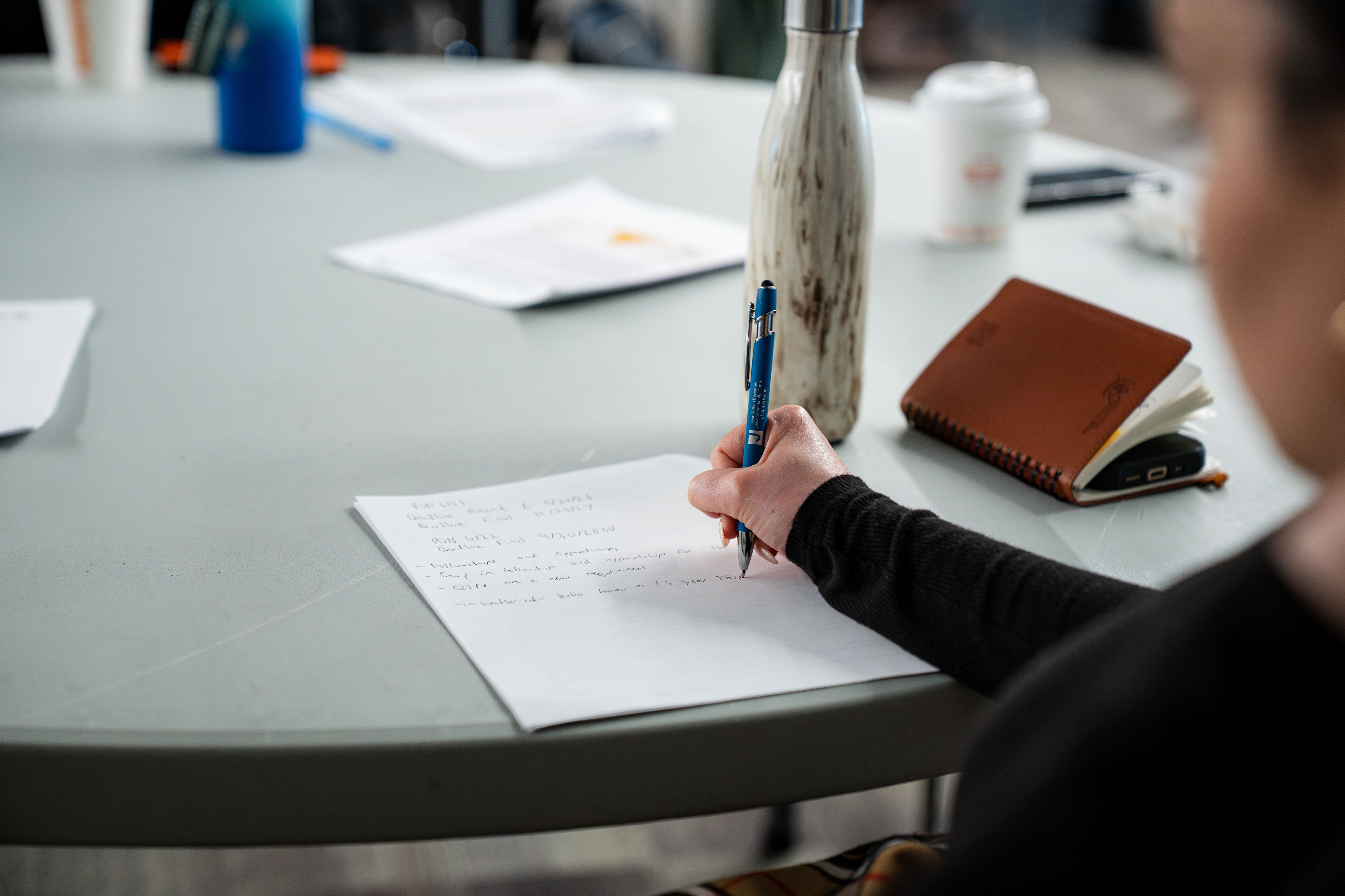 A woman writing notes on a piece of paper at a circular table with other meeting papers.