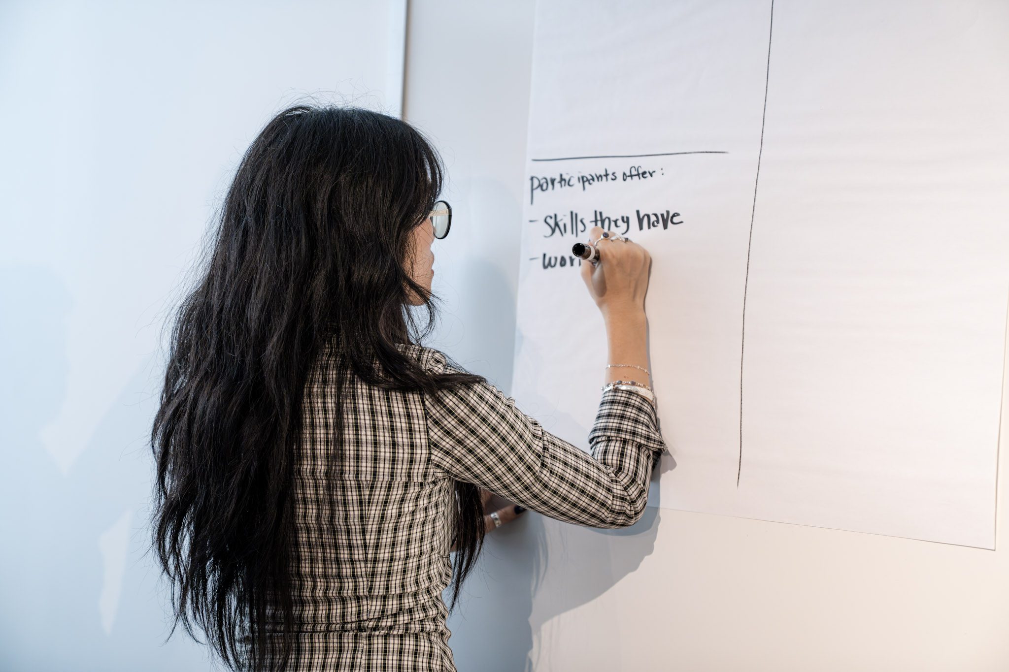 A photo of a woman with long black hair wearing a black and white plaid shirt writing on a large post-it note affixed to an office wall.